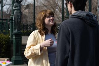 Actor in a pale yellow shirt smiling mid-conversation, the park statue and bandstand behind her