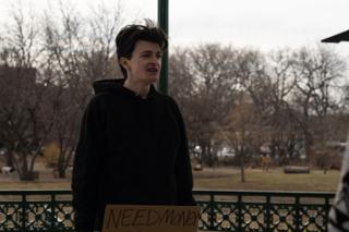 Actor in a black hoodie speaking in the park bandstand, a hand-lettered cardboard sign held at his waist