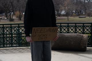 Close on the hand-lettered cardboard sign reading NEED MONEY FOR SURGERY — DYING, held against the bandstand railing