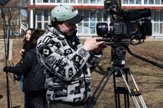 Camera operator in a patterned fleece framing up on the tripod, the schoolyard windows behind him
