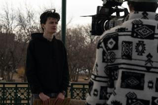 Operator in a patterned fleece shooting handheld while the actor stands with his sign in frame