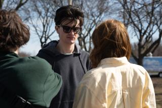 The two leads talking between takes, the actor in sunglasses listening with the park behind