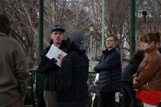 The director in a flat cap talking the scene through with the day's pages in hand, the company gathered at the bandstand