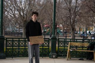 The actor standing square to camera with the NEED MONEY FOR SURGERY sign, the playground behind the railing