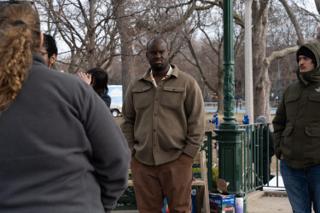 A man in a tan shirt jacket standing in the scene while the company watch from the foreground