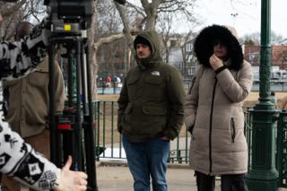 Two of the background cast waiting in winter coats behind the camera tripod
