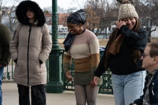 The background cast laughing together at the railing, the seated actor in the corner of frame
