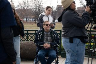 An actor seated in a wheelchair at the bandstand as the operator moves in to film him