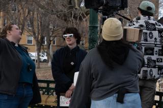 Cast and crew grouped around the camera between takes, the actor holding his sign at the railing