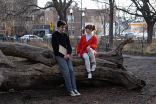 The two leads sitting apart on the fallen log with the cardboard sign between them