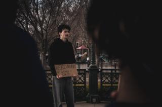 The actor and his sign seen between two out-of-focus onlookers watching from the foreground
