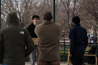 A passer-by in a fleece stopping in front of the actor and his sign while others look on