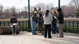 The crew in a huddle around the camera on the bandstand, a cast member waiting with his pages on the log