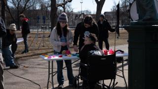Paper flowers and coloured paper spread across the prop table, a crew member in a wheelchair folding one with two of the company alongside