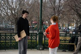 A woman in a red sweater standing across from the actor and his sign beside the park lamppost