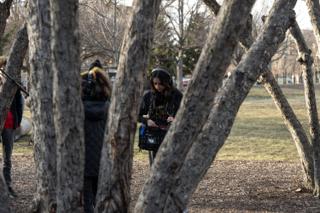 The sound recordist working the mixer bag seen between the tree trunks, headphones on and the boom operator waiting behind her