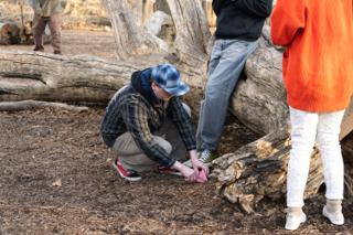 A crew member crouching to set the pink paper flower on the ground at the actor's feet, the leads waiting either side