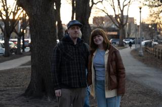 The two leads standing together on the park path at sunset, streetlights coming on along the walk