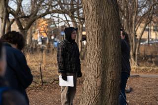 A crew member in a hooded plaid shirt holding a script beside the trunk of a large tree
