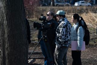 The camera operator lining up a shot on sticks at the base of a tree, two of the crew watching from behind