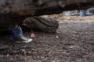 Low on the ground beside the log — a dropped pink paper flower at the actor's feet