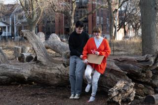 The two leads leaning against the fallen log, her hands cupped around the pink paper flower over the sign