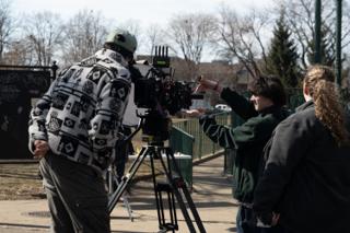 A crew member reaching in to the camera as the operator lines up a shot, the chalkboard and green railings behind