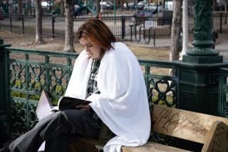 Actor wrapped in a white blanket on a park bench, reading between setups