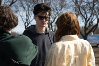 The actor in sunglasses listening to the actress, the bare park and an ice cream truck behind them
