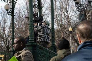The camera operator up inside the bandstand ironwork with the camera on his shoulder, three of the crew watching from below