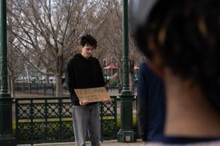 The actor looking down at his hand-lettered cardboard sign inside the bandstand, a crew member close in the foreground