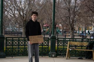 The actor alone in the middle of the bandstand with his sign held at his waist, the playground and bare trees behind him