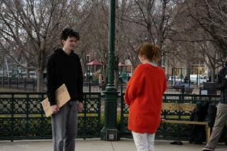 The actor turning with his sign towards the woman in the red cardigan, the two of them framed by the green bandstand posts