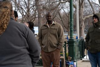 An actor in a brown overshirt standing by for the take, crew shoulders close in the foreground and the bandstand ironwork behind