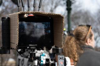 A cardboard sunshade taped around the on-camera monitor to cut the glare, the operator watching behind it
