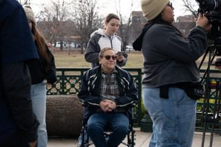 The older actor waiting in his wheelchair while his hair is touched up, the operator stood over the A-camera at the right