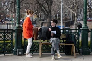 The seated actor going through his pages in sunglasses while the woman in the red cardigan stands over him with a coffee, coats piled on the bandstand bench