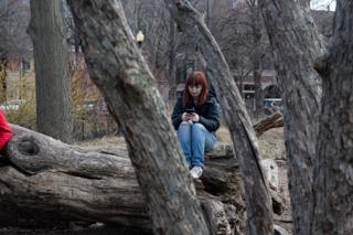 A crew member alone on the fallen log with her phone, framed by the bare branches