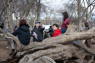 The director pointing out the blocking to the cast gathered along the fallen log, pages in his hand