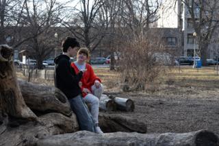 The two leads sitting together on the fallen log mid-scene, the park and apartment blocks behind them