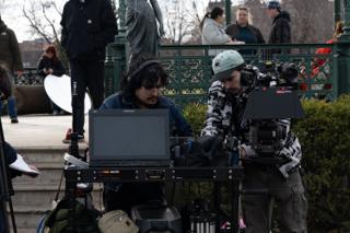 Two crew bent over the monitor cart beneath the park statue, the camera built up alongside