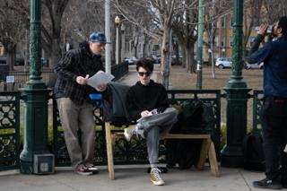 Director in a flat cap holding script pages, talking to the actor sitting on the bandstand bench