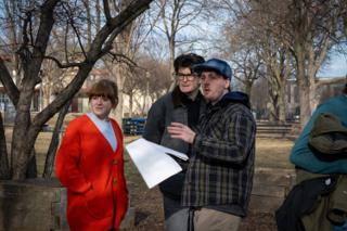 Director in a blue flat cap talking a scene through with two of the cast, script pages in his hand