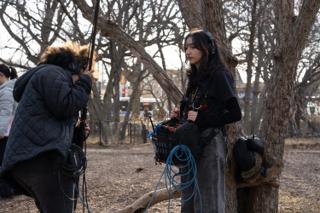 Boom operator leaning in with the pole while the recordist works the mixer bag against a tree, blue cable looped at her feet