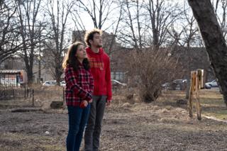 Two of the cast standing together in the dry garden, both looking up and off past the camera