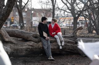 The leads sitting together on the fallen log mid-scene, script pages held out of focus in the foreground