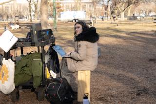 Crew member in headphones working a laptop on her knees beside the monitor cart, wrapped up against the cold