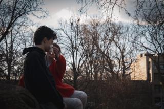 The two leads close together on the log at dusk, a folded paper flower held up between them