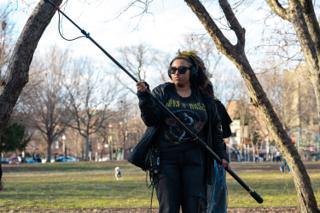 Boom operator carrying the pole across the park, headphones on and cable trailing behind