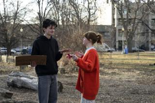 The actress gesturing with a red paper flower as the actor listens, his cardboard sign lowered at his side